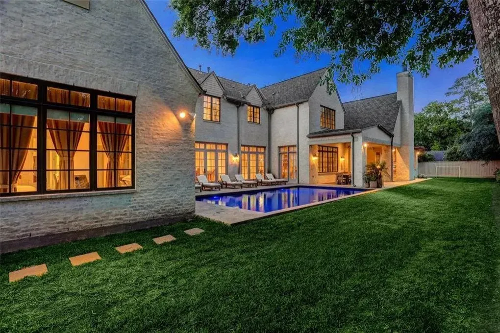 Backyard of a large house with a pool, green lawn, and trees at dusk. Exterior lights illuminate the building.
