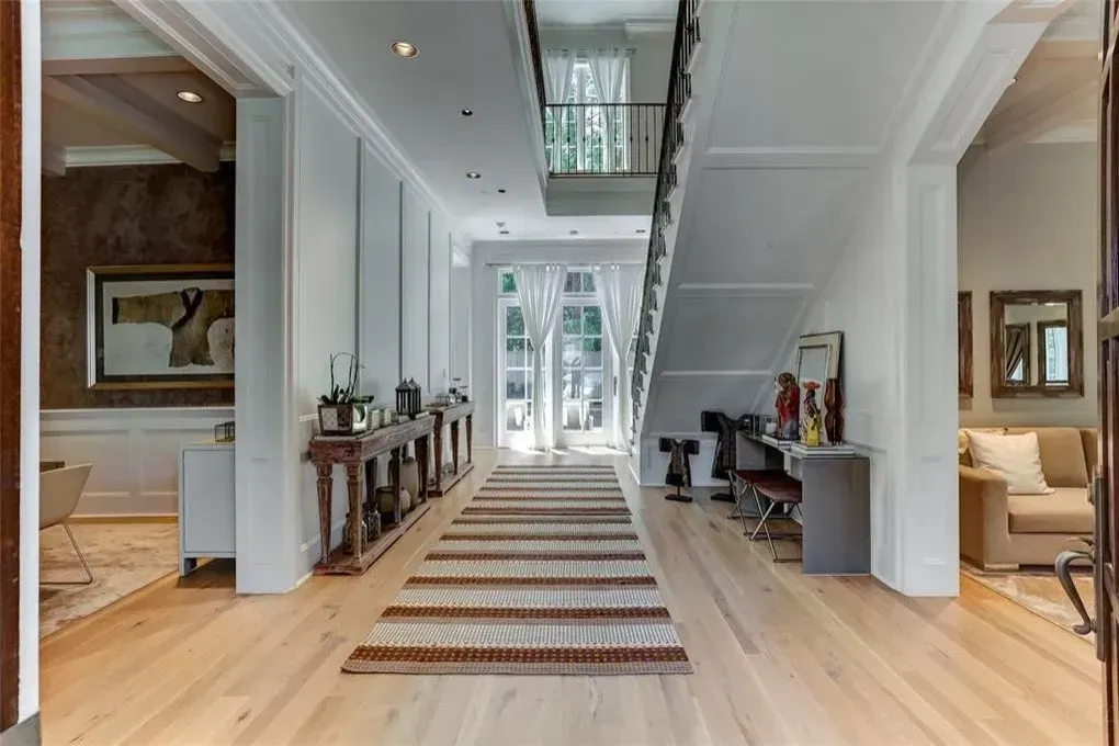 Hallway with wooden floor, staircase, and long rug. Sunlight streams through the doors at the end.
