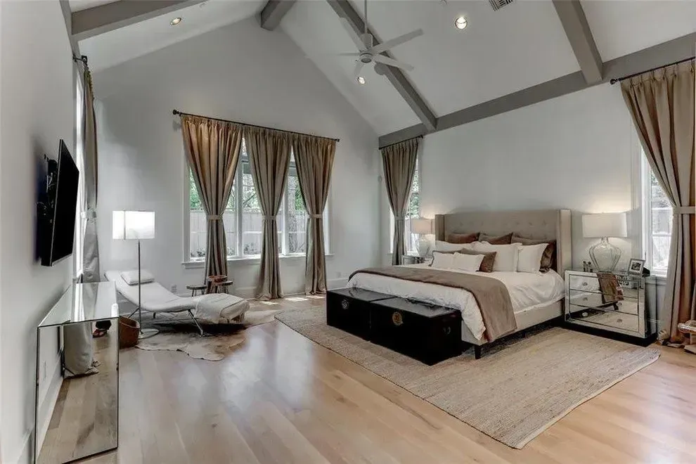 Spacious bedroom with vaulted ceiling, large windows, neutral tones, and a bed with a trunk at the foot.