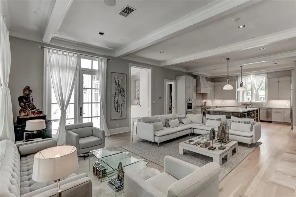 Spacious, bright living room with white sofas, glass tables, and kitchen in the background.