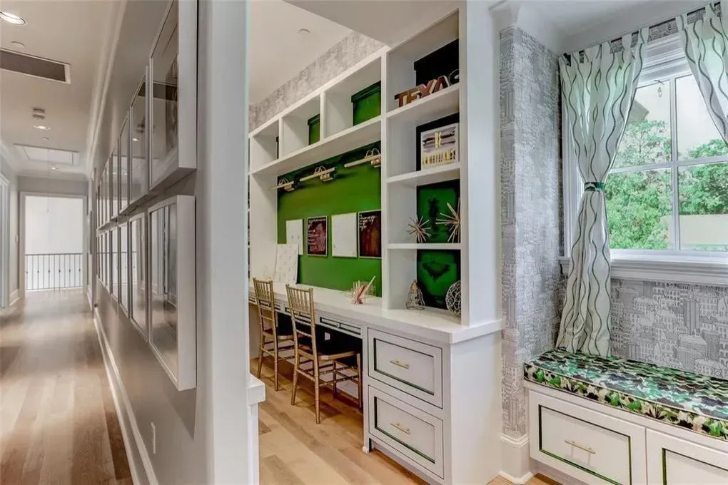 Built-in white desk with green accents and bookshelves in a hallway, near a window with patterned curtains.
