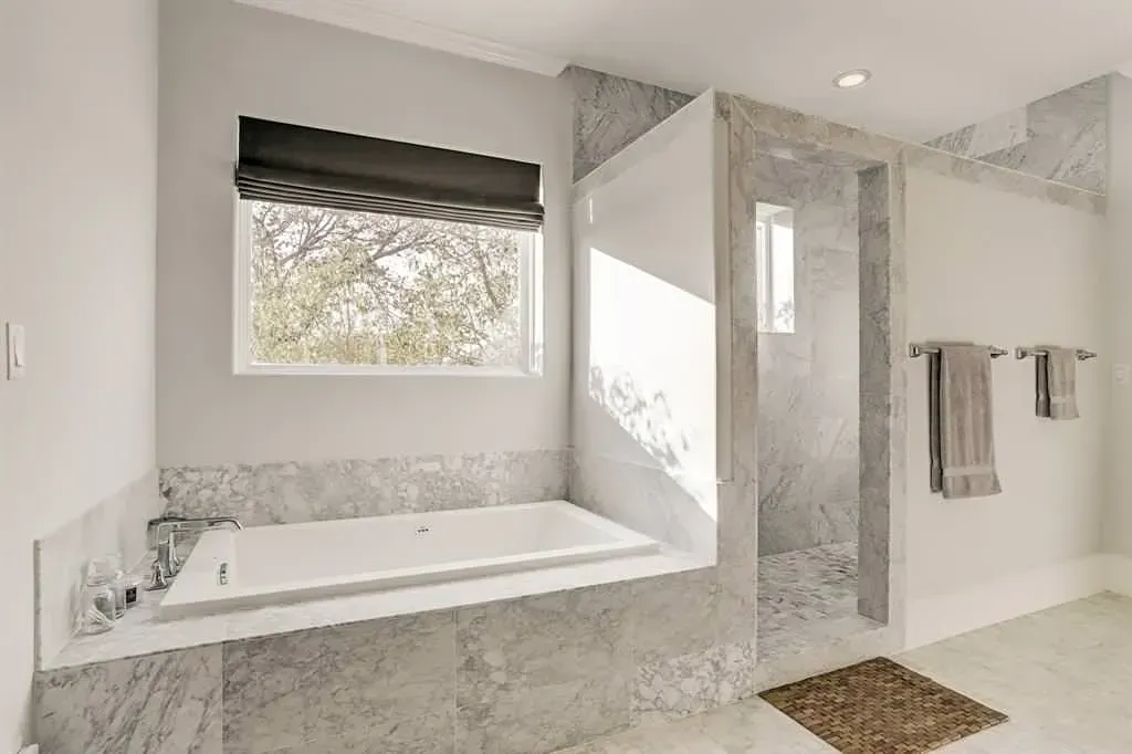 Modern bathroom with a tub, shower, and window. Gray stone tile, gray blinds, and two towel racks.