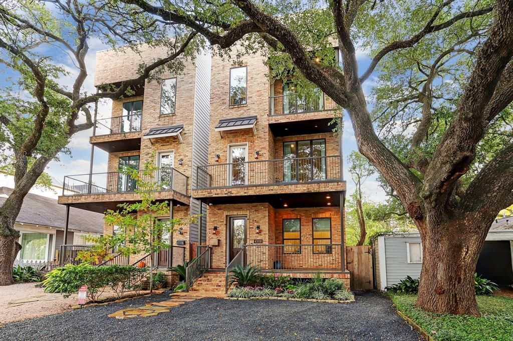 Brick multi-story house with balconies, mature trees, and gravel driveway.
