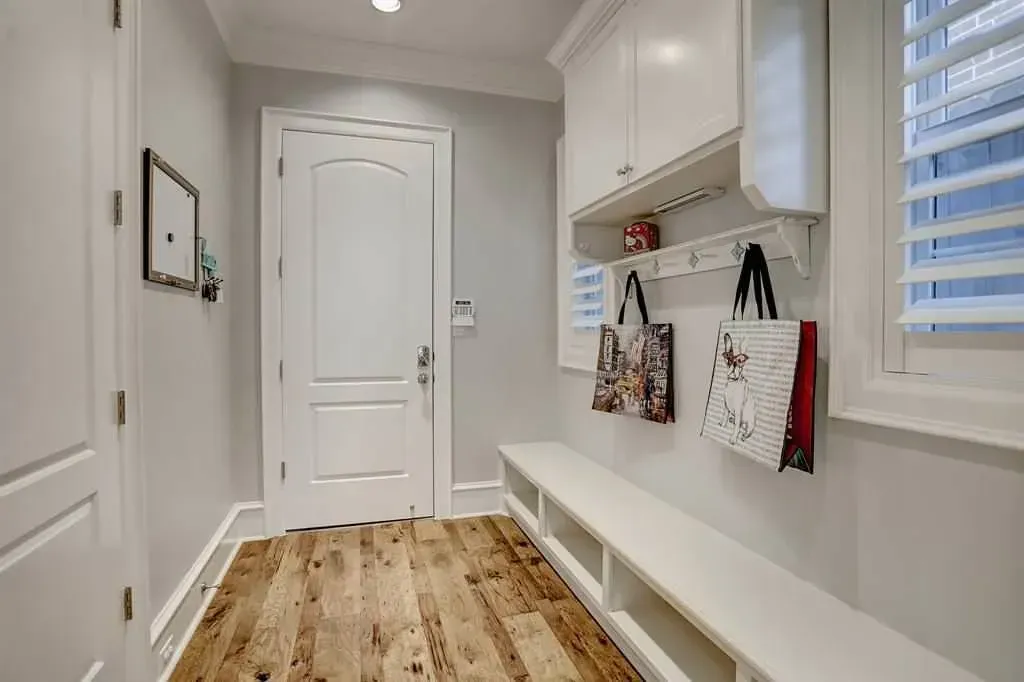 A bright entryway with a bench, storage, and a white door. Wooden floor and light gray walls.