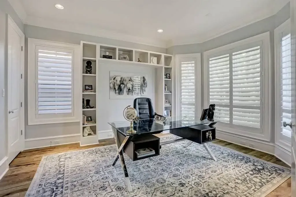 Home office with built-in bookshelves, large windows with shutters, glass desk, patterned rug, and a leather office chair.