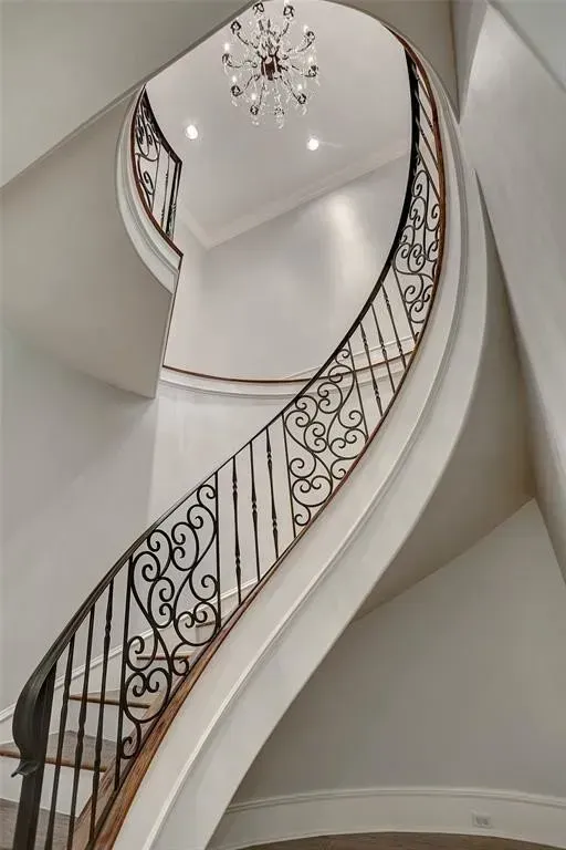 Curving staircase with ornate black wrought-iron railing against light gray walls; chandelier hangs overhead.