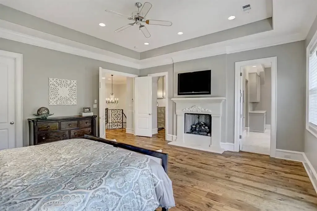 Bedroom with fireplace, hardwood floors, and light gray walls. A bed is in the foreground, and there are open doorways.