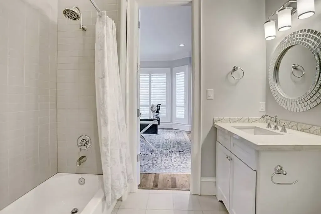 Bathroom with white tile, bathtub, shower curtain, vanity, mirror, and doorway to a living area.