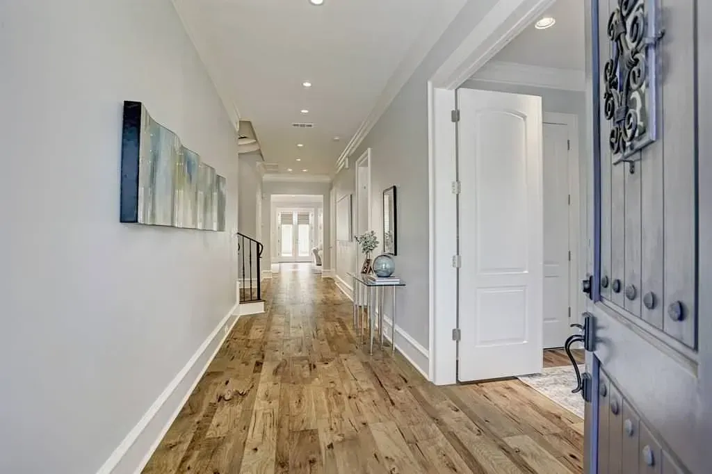 Long hallway with light wood floors, white walls, artwork, and a partly open door.
