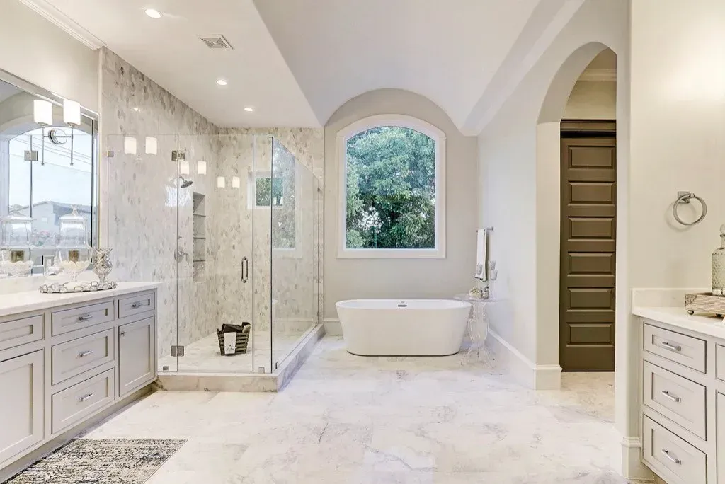 Elegant bathroom with marble floors, glass shower, freestanding tub, and a large arched window.