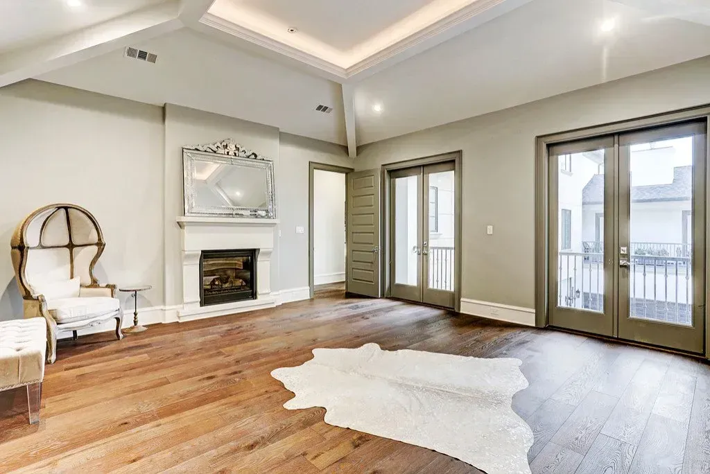 Spacious bedroom with wooden floors, fireplace, and French doors leading to a balcony. A cowhide rug is on the floor.