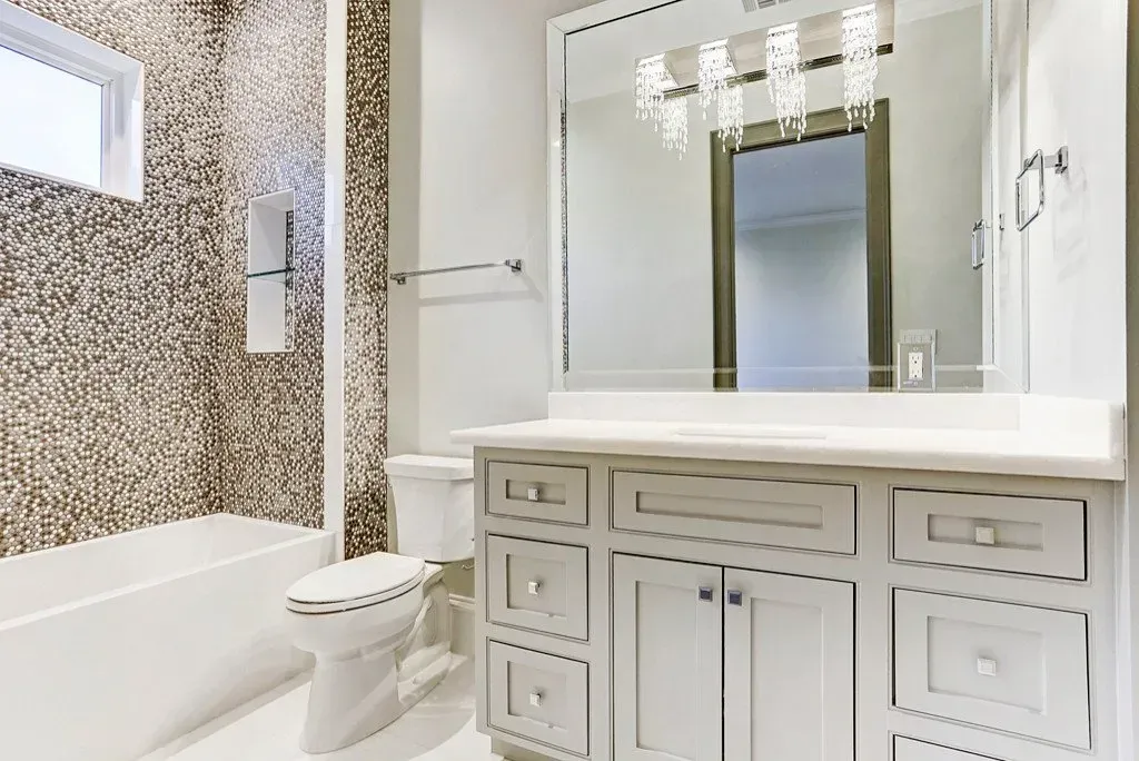 Bathroom with mosaic-tiled shower, white vanity, toilet, mirror, and bright lighting.
