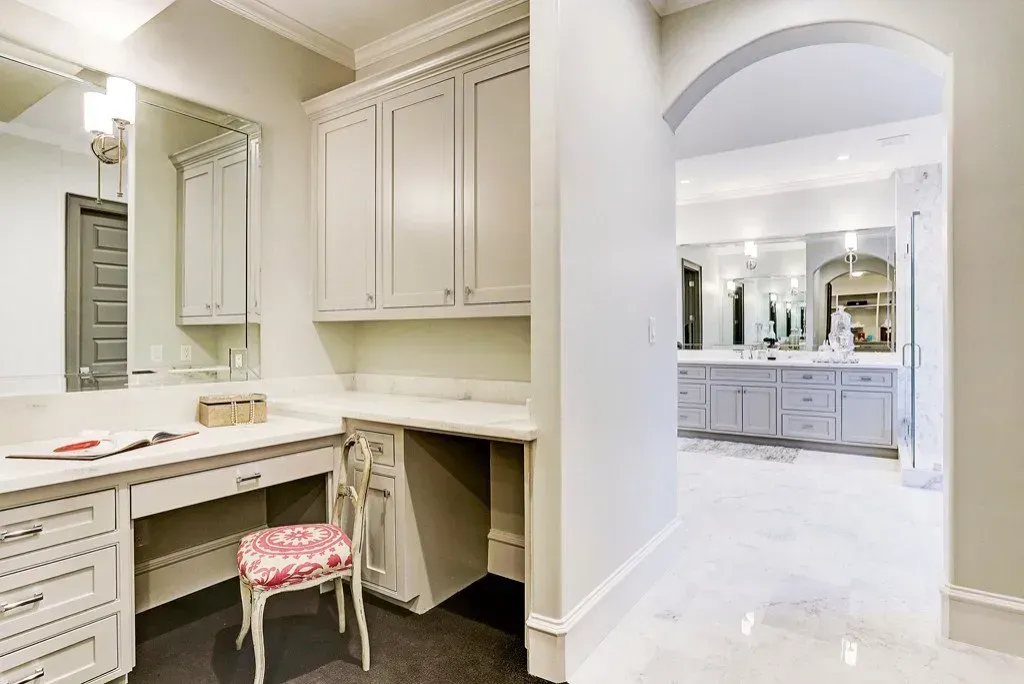 Vanity area with a desk, cabinets, and a doorway leading to a bathroom with double sinks.