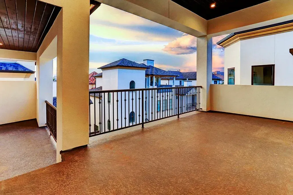 Open air balcony with black railing, brown speckled floor, and view of white buildings under a sunset sky.