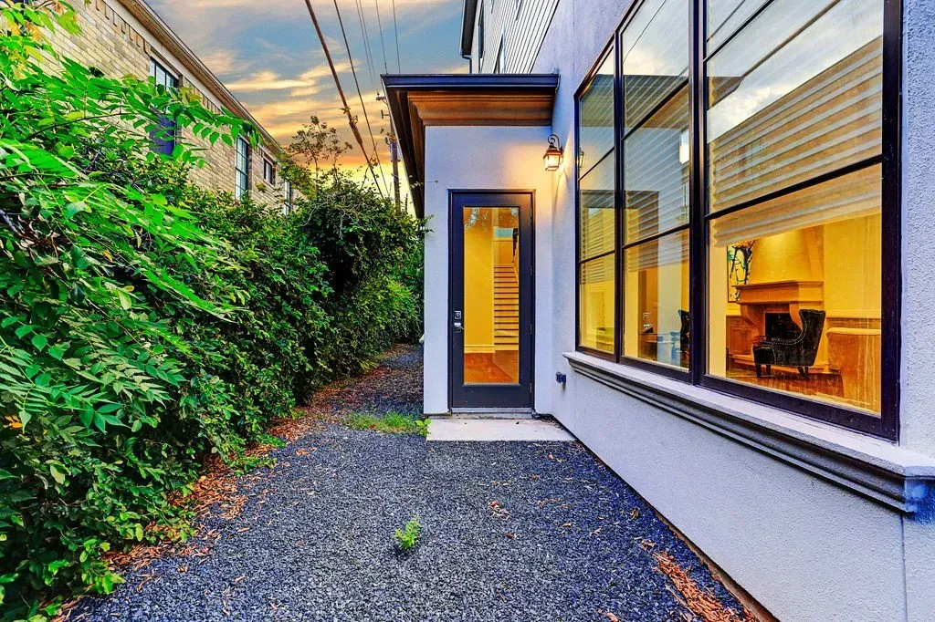 Exterior of a modern home with black gravel ground, shrubs, and large windows reflecting a warm interior.