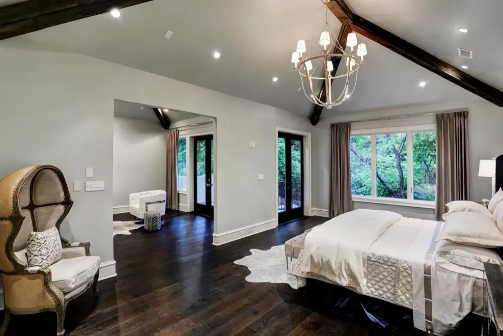 Spacious bedroom with dark wood floor, large window, chandelier, and a chair.