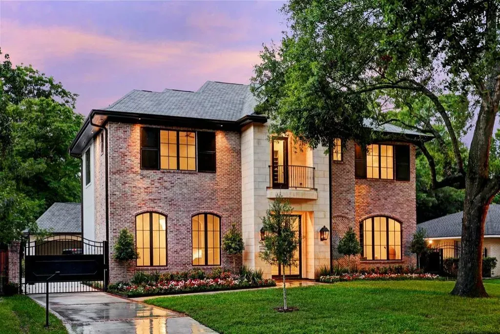Two-story brick house with dark shutters and arched windows at dusk, lush green lawn, and a driveway.