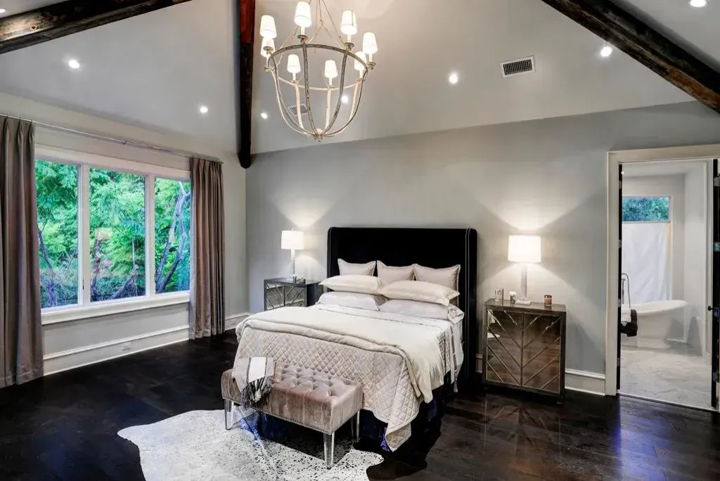 Bedroom with dark hardwood floors, large window, bed, and chandelier. Adjacent bathroom visible.