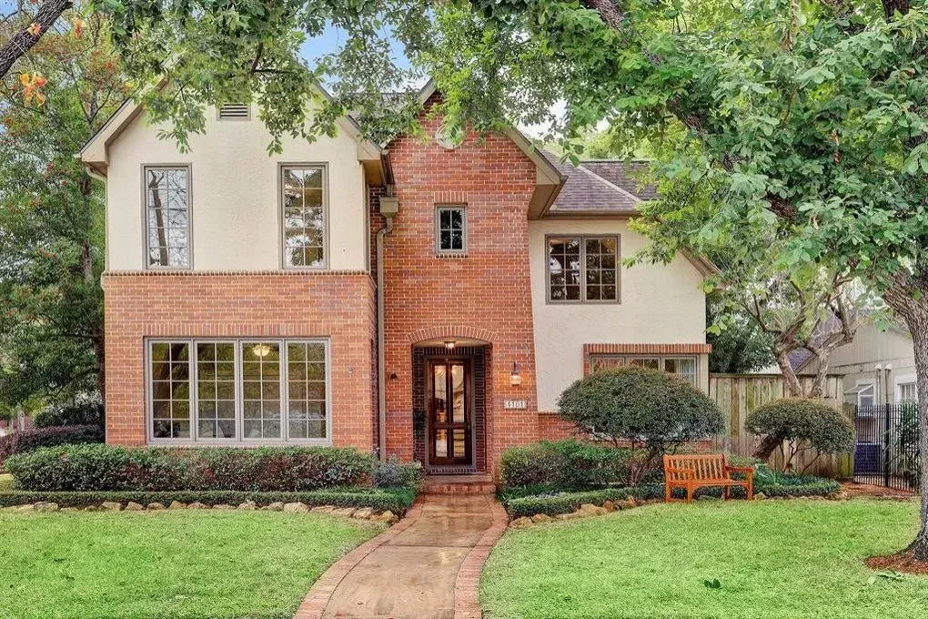 Two-story brick and stucco house with a path leading to the front door, set amidst green lawn and trees.