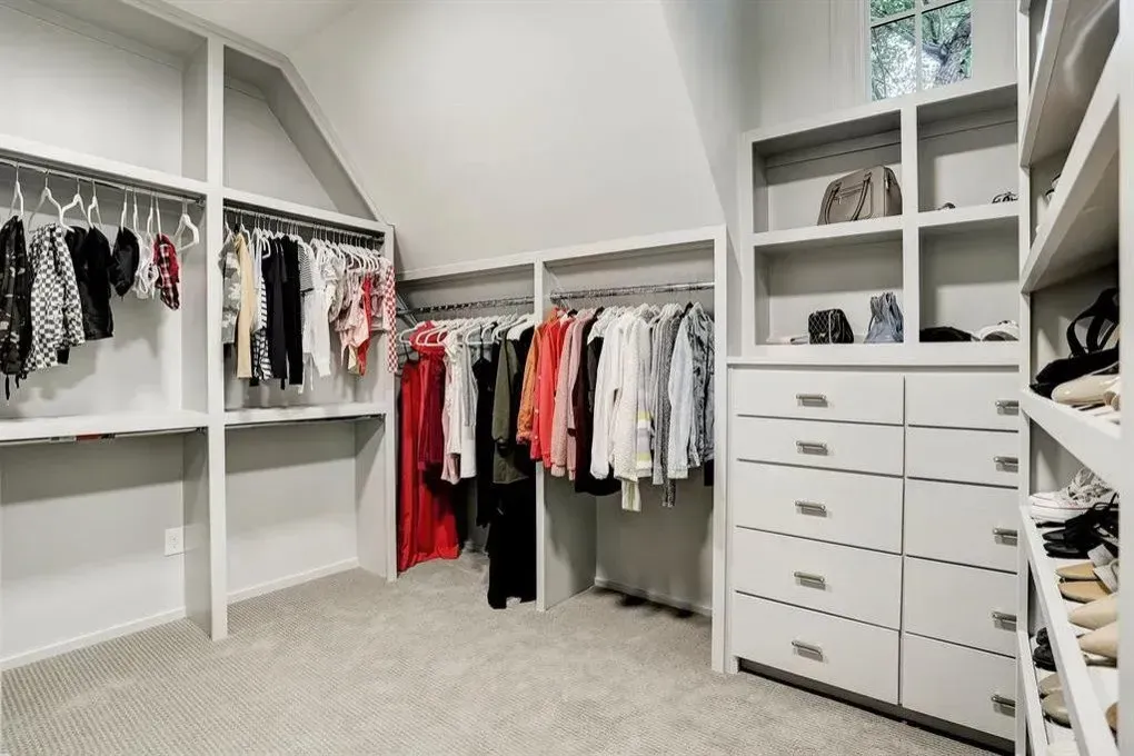 Walk-in closet with white shelves and rods, filled with clothes. Gray carpet, angled ceiling, bright.