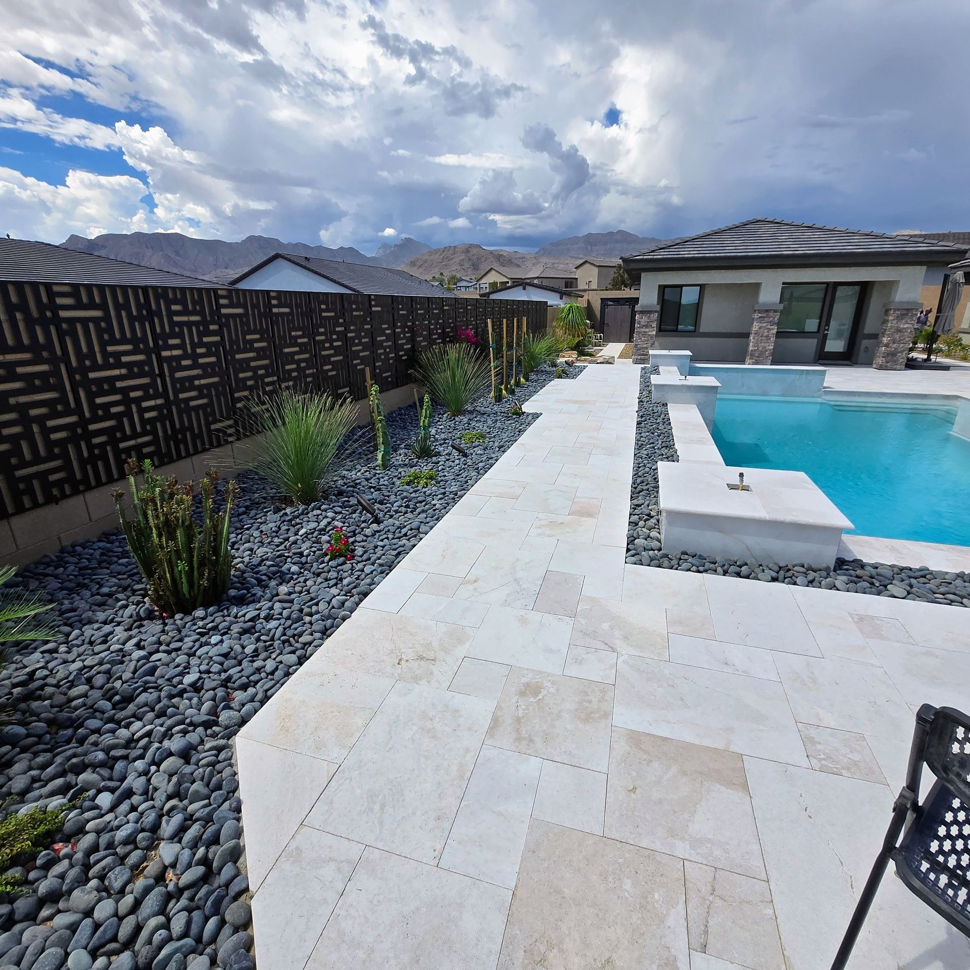 Backyard with a pool, patio, decorative black rocks, and a patterned fence. Cloudy sky and mountains in the background.