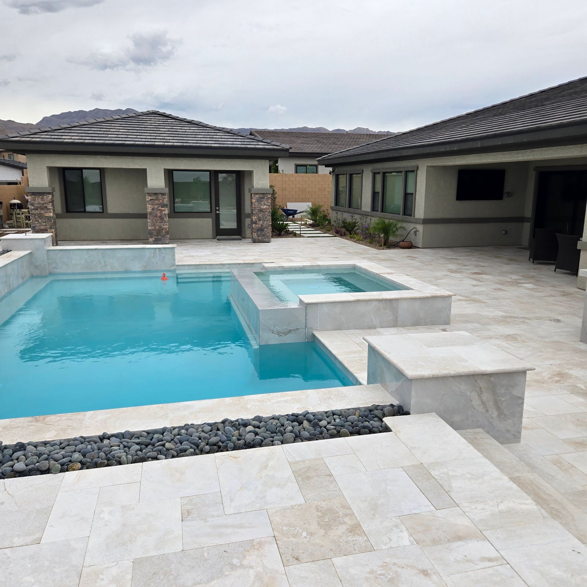 Backyard with a pool, spa, and patio. Light blue water, gray stone, and a house under a cloudy sky.