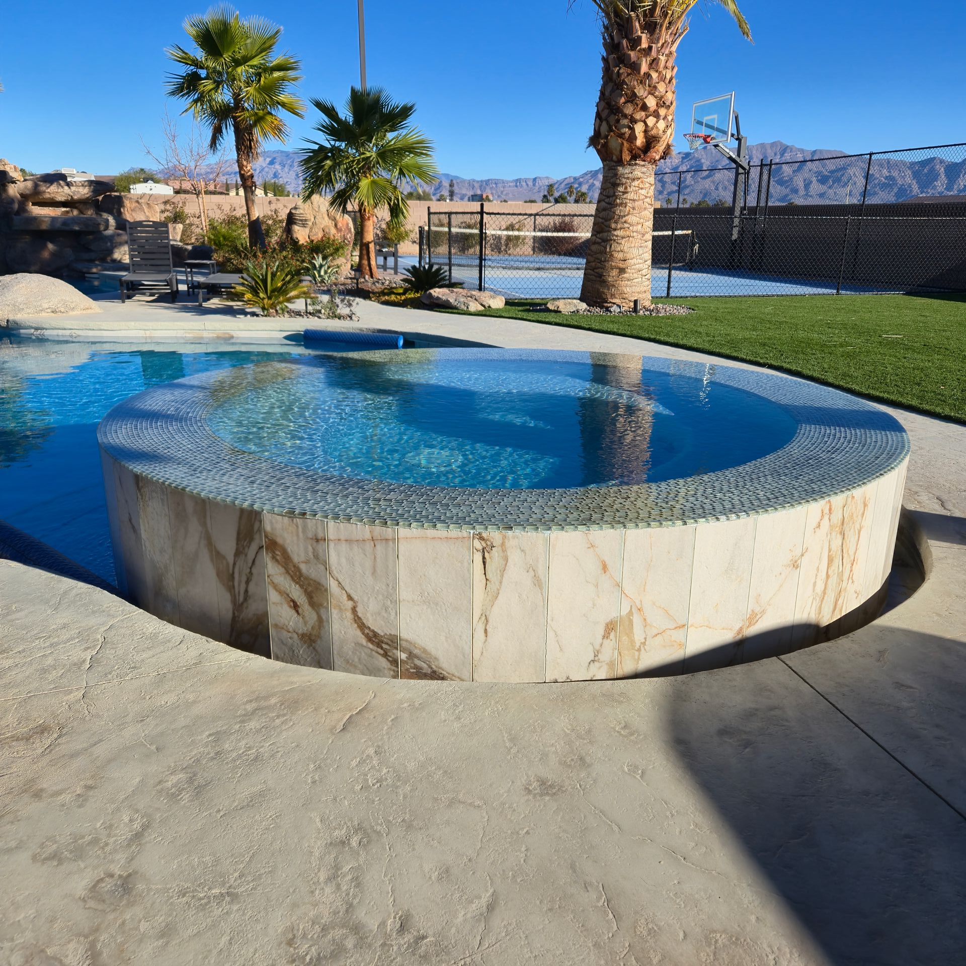 A luxurious pool and spa area with a stone-clad hot tub. Blue water, palm trees, and artificial grass.