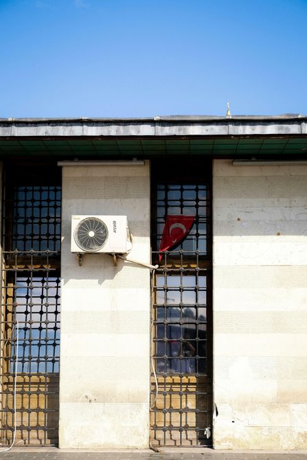 White brick building facade with barred windows, a wall-mounted air conditioner, and a red cloth behind one window.