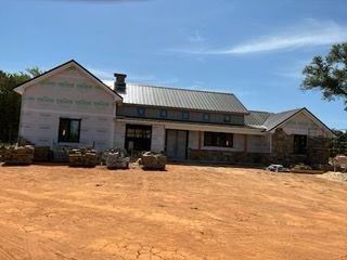House under construction with stone accents, metal roof, and surrounding dirt ground under a blue sky.