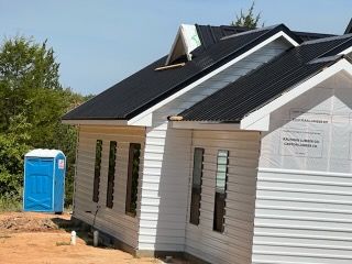 House under construction with black roof, white siding, and blue portable toilet nearby.