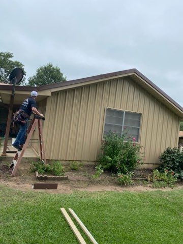 Person on ladder repairing roof siding on a light brown house with brown trim. Green lawn and bushes in yard.
