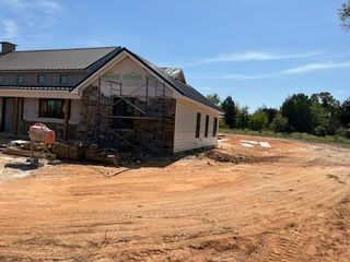 Construction site with house under development; stone and siding exterior, scaffolding, dirt lot, blue sky.