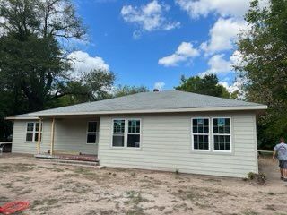 Beige-sided house under construction with multiple windows. A person walks by on a dirt yard under a cloudy blue sky.