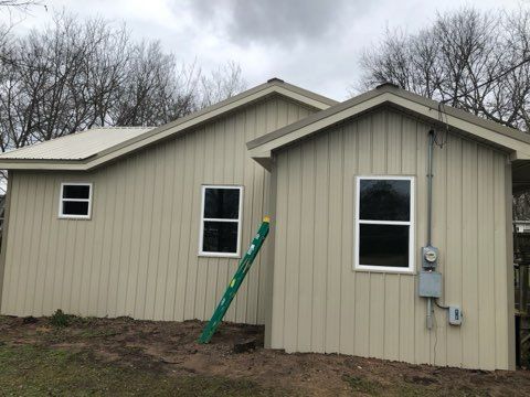 Tan-sided buildings with white-framed windows, a green ladder, and an electrical box. Overcast sky and bare trees.