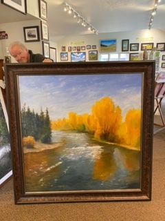 A man is holding a framed painting of a river surrounded by trees.