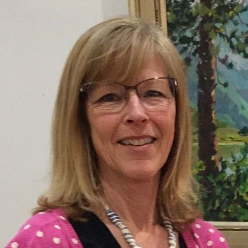 A woman wearing glasses and a necklace is smiling in front of a painting.