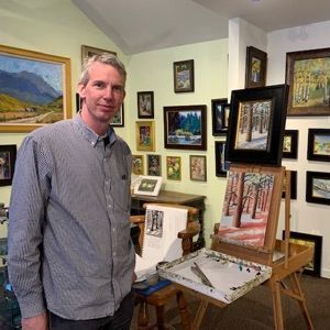 A man is standing in front of an easel in a room with paintings on the wall.