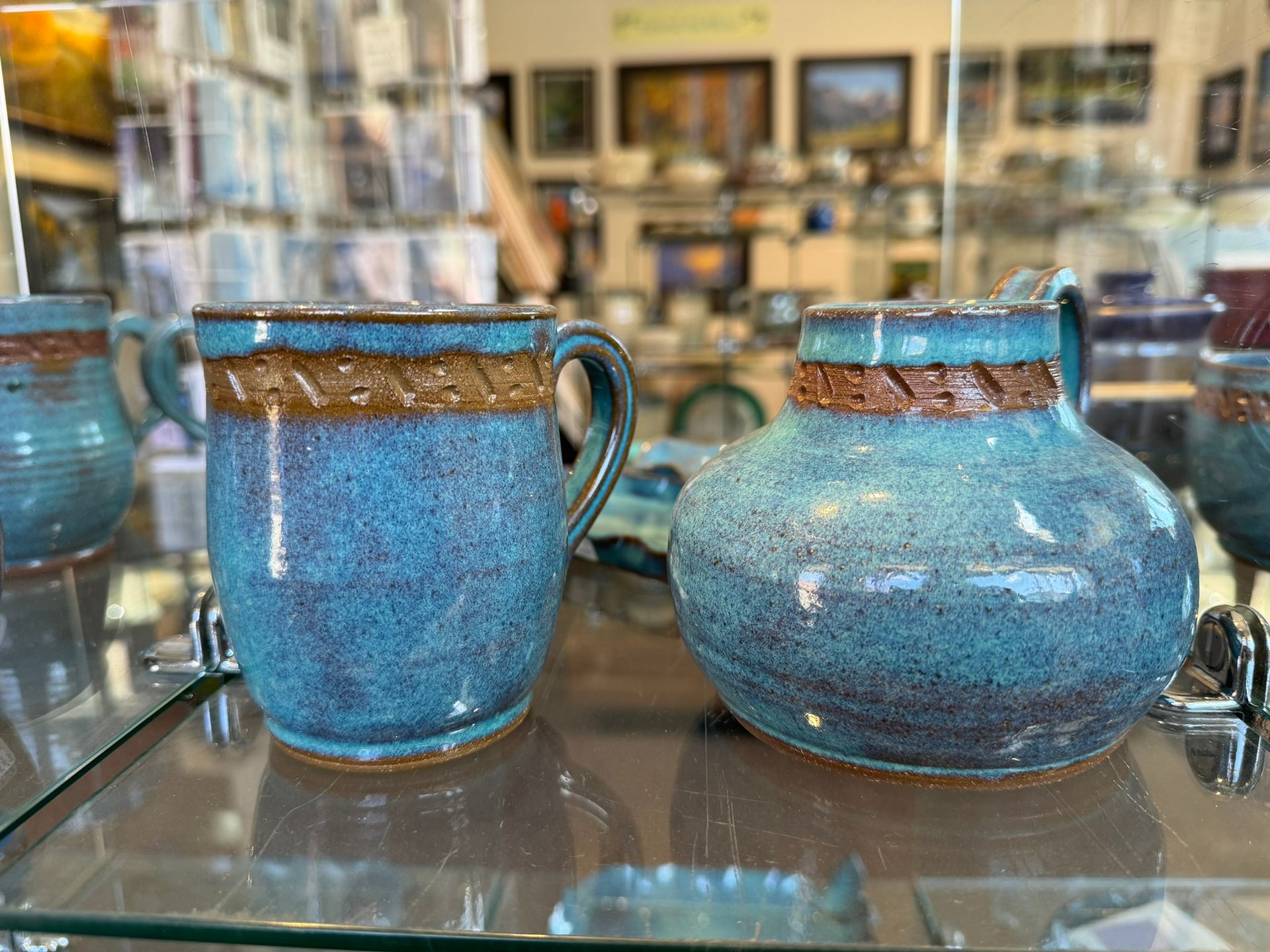 Two blue mugs and a vase are sitting on a glass shelf in a store.