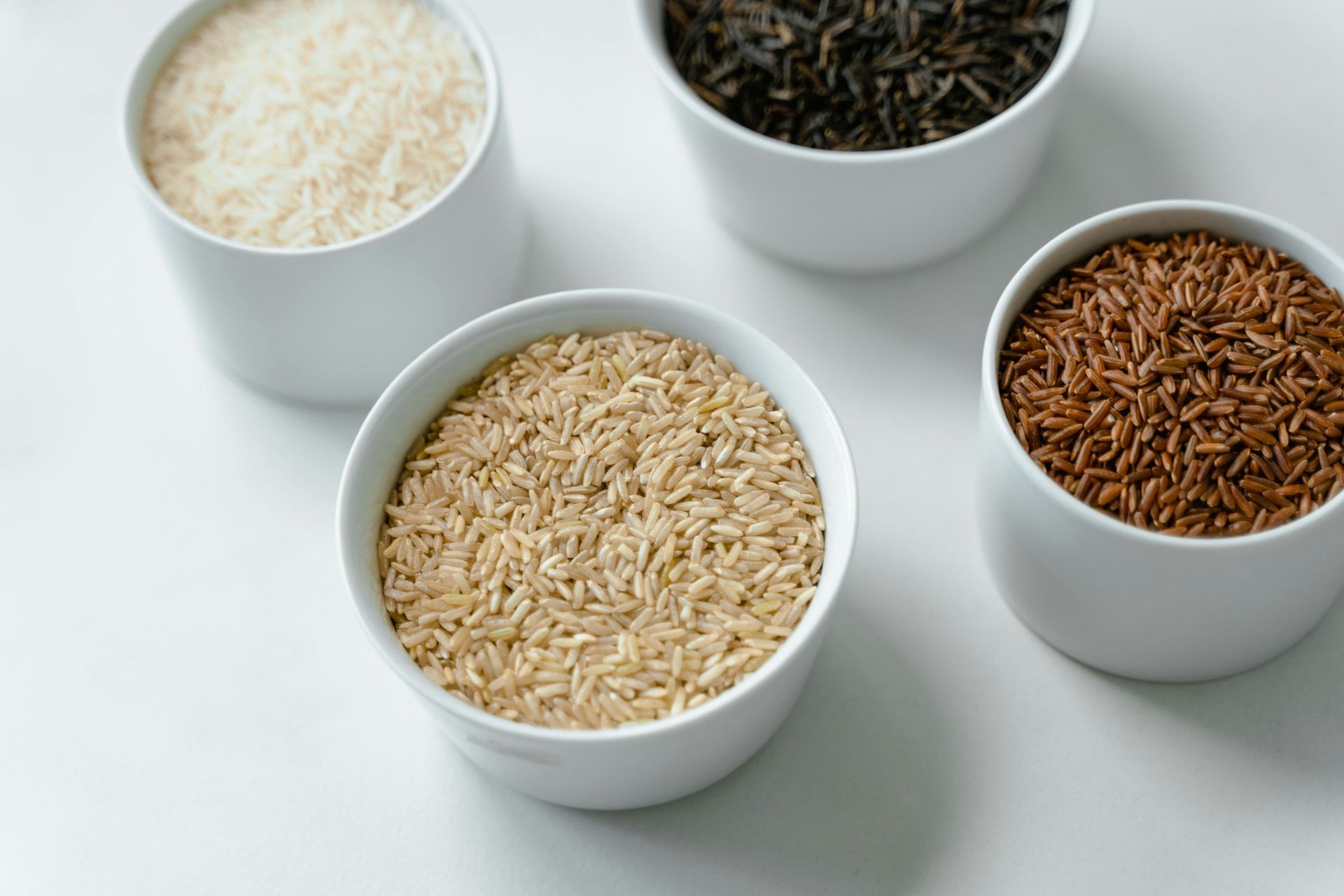 Four white bowls of assorted rice grains on a light surface.