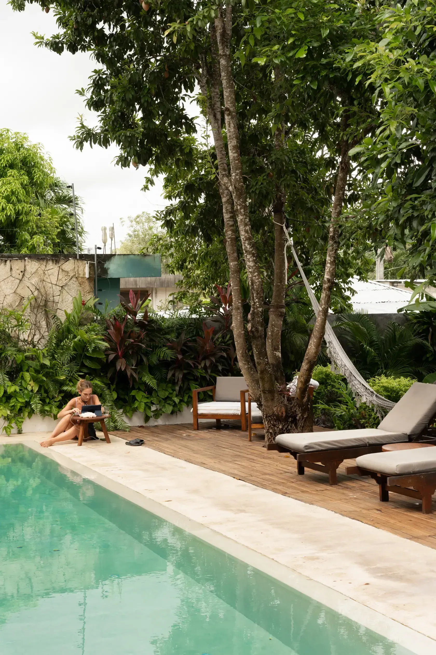 Mujer leyendo junto a la piscina, con árboles, tumbonas y exuberante follaje verde.