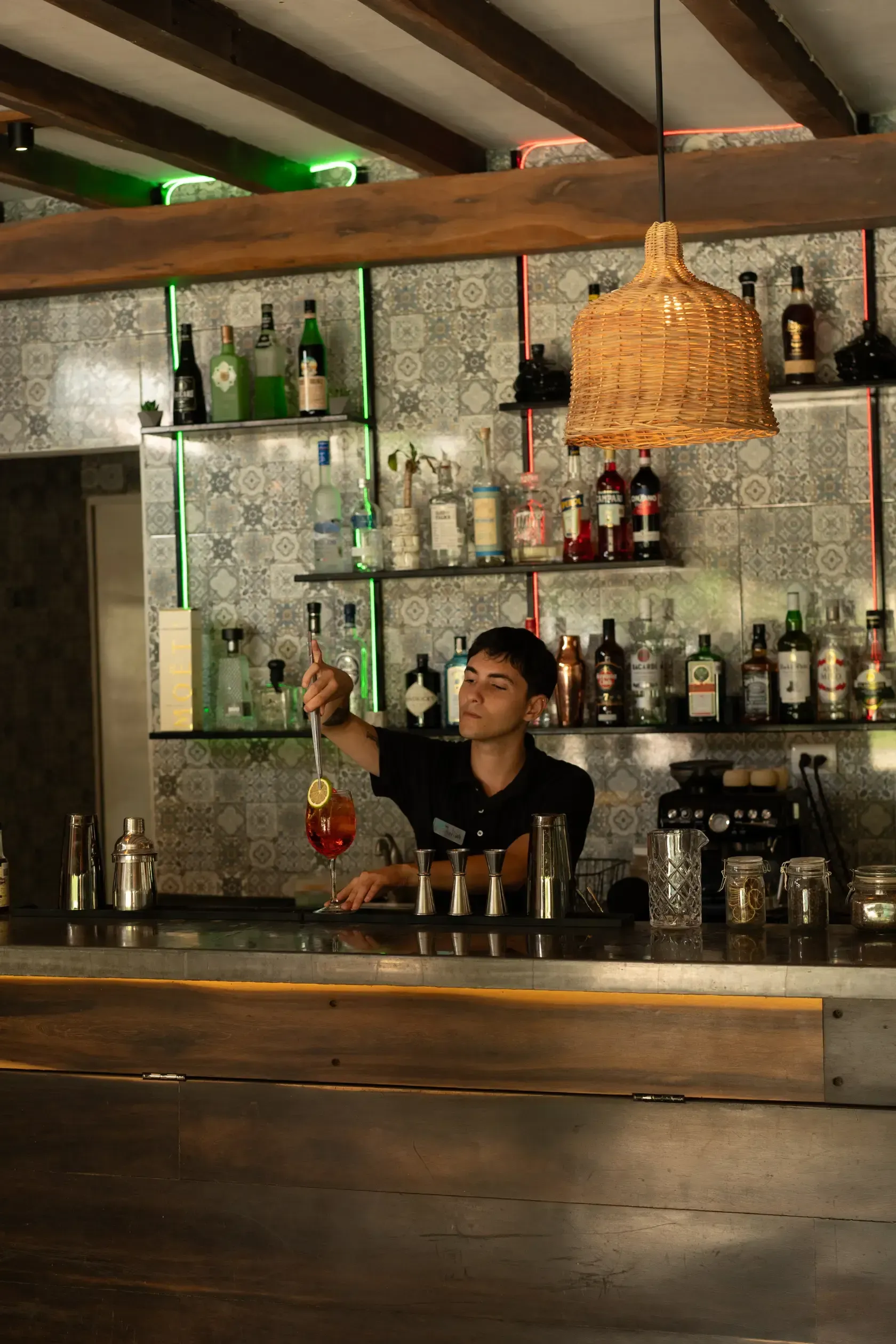 Camarero con camisa negra revolviendo un cóctel en una barra con botellas, estantes y una lámpara de mimbre.