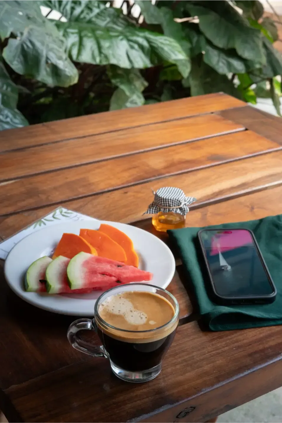 Desayuno: fruta en rodajas, café, miel y teléfono sobre una mesa de madera con un fondo de hojas verdes.