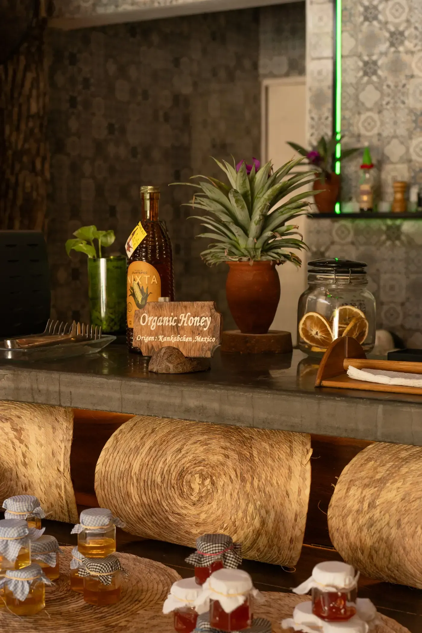 Barra de bar con bebidas y plantas decorativas. Tonos cálidos con frascos, botellas y una piña.