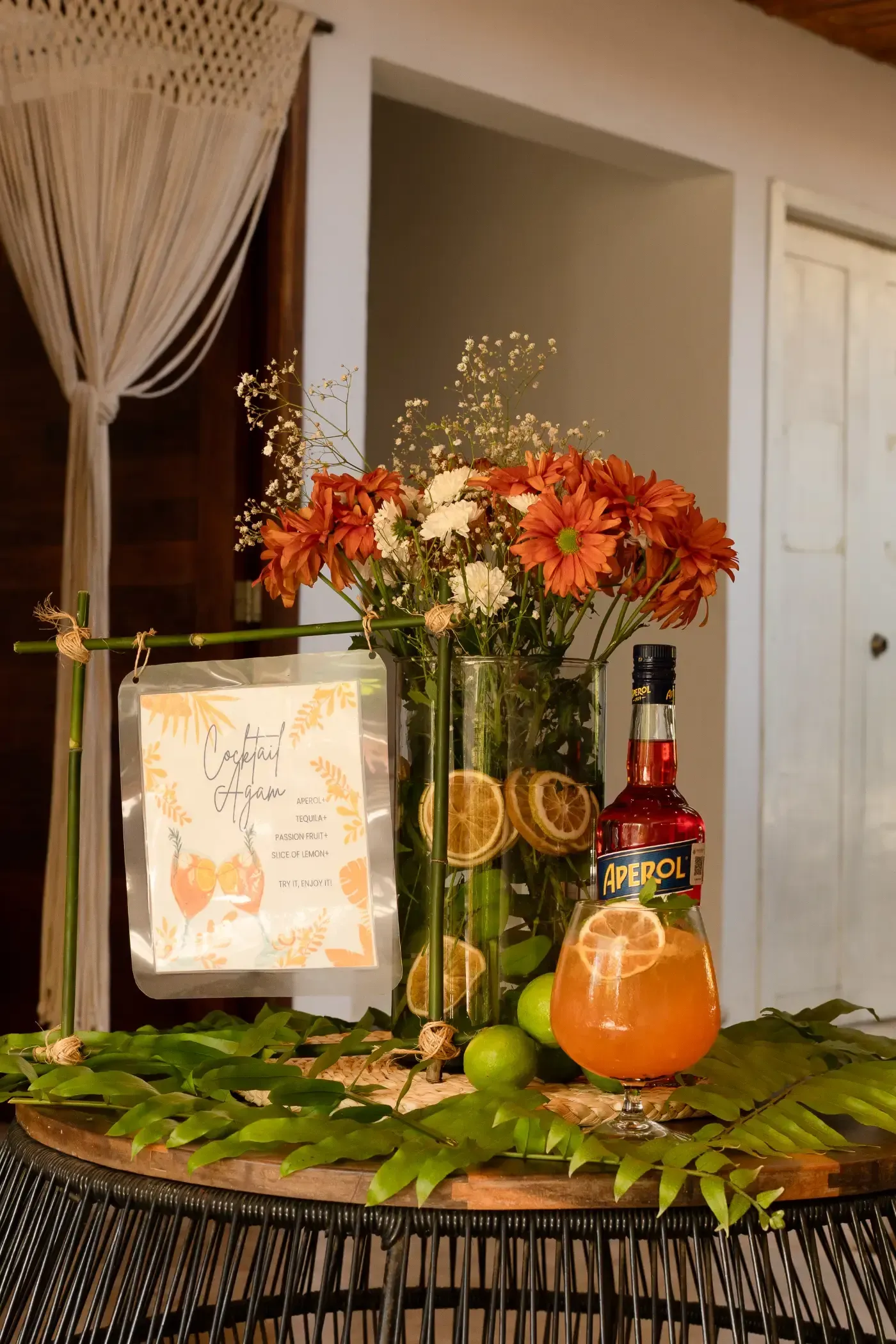 Montaje de bar de cócteles con flores, frutas, un cartel y una botella de licor, al aire libre.