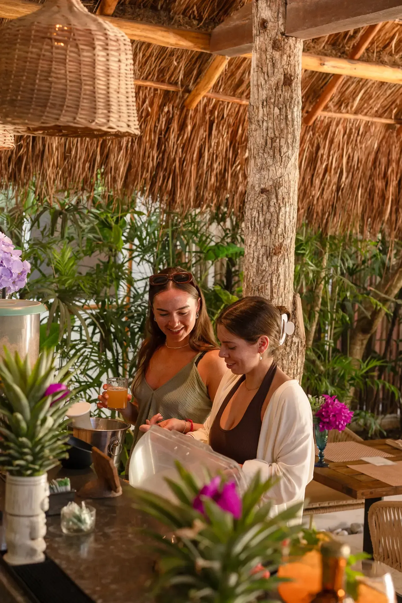 Dos mujeres en un bar en un entorno tropical, una preparando una bebida y la otra mirando.