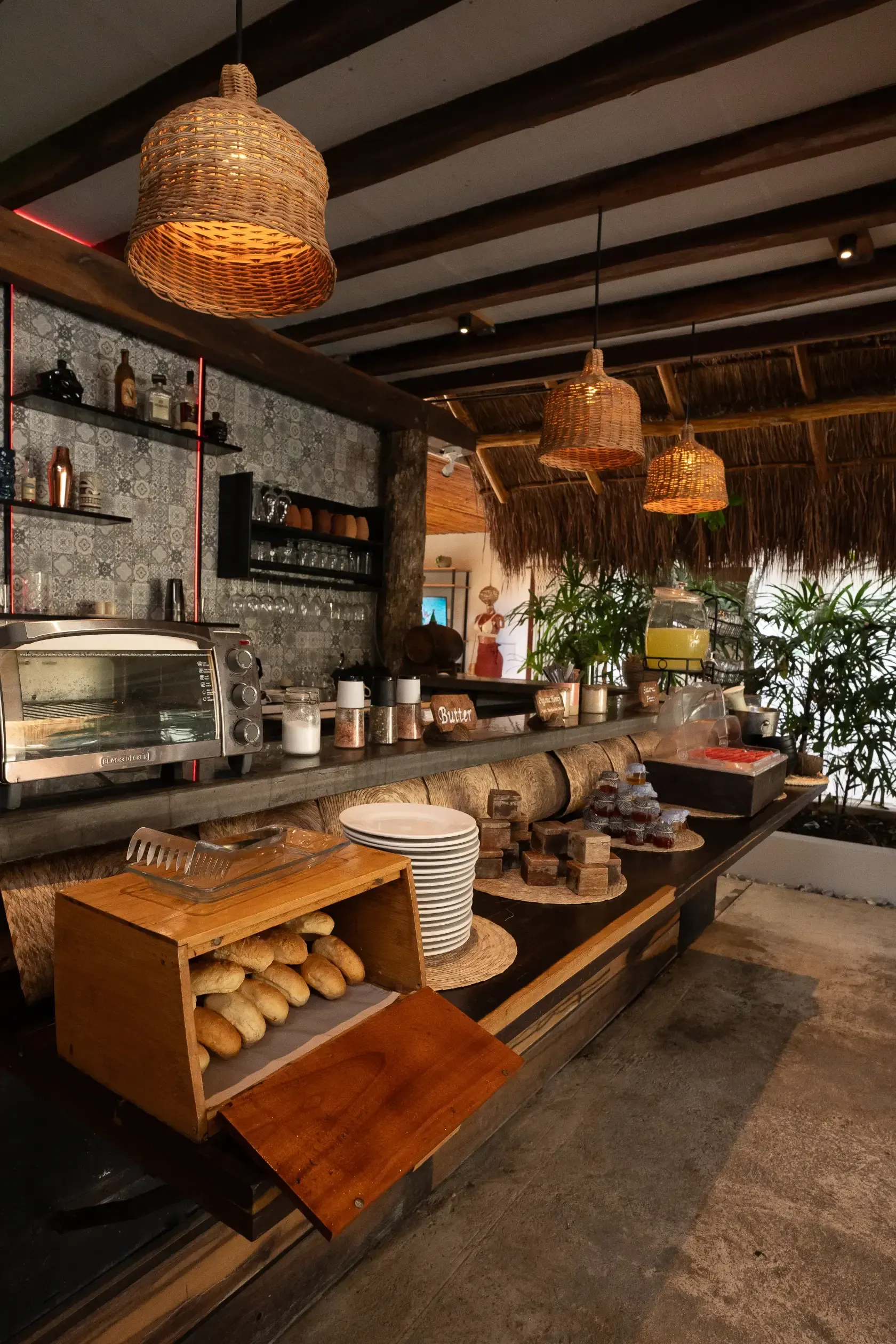 Mostrador de restaurante con productos horneados, bebidas y lámparas colgantes tejidas; entorno tropical.