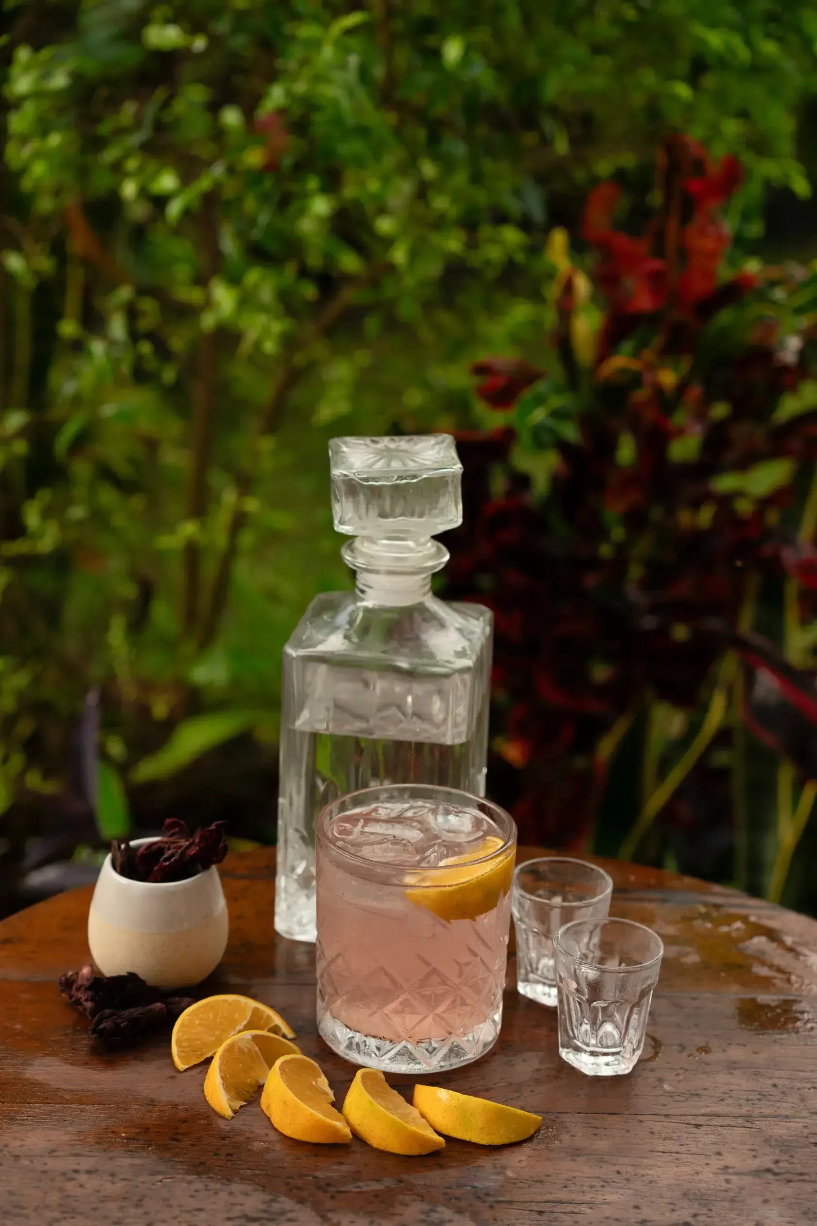 Un cóctel rosa con rodajas de limón, una botella de licor y vasos de chupito en una mesa de madera al aire libre.
