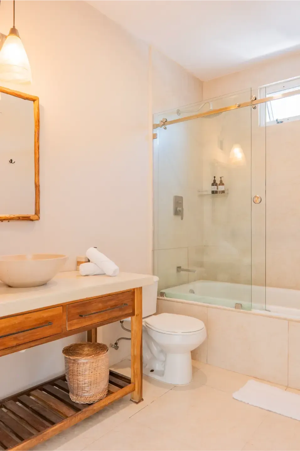 Baño con tocador de madera, lavabo de piedra, ducha de cristal y azulejos blancos.