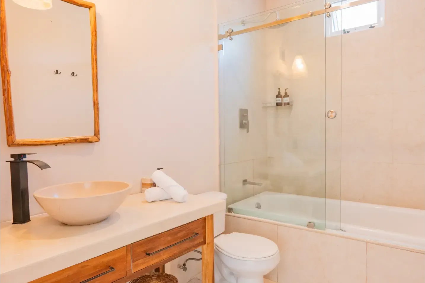 Baño con paredes beige, tocador de madera, lavabo, espejo y ducha detrás de una puerta de vidrio.