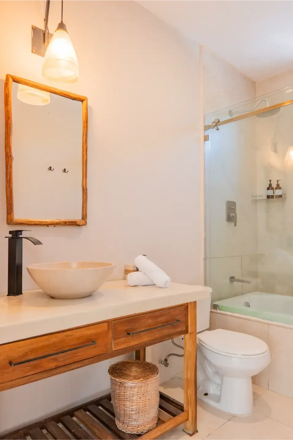 Baño con tocador de madera, lavabo sobre encimera, inodoro y ducha de cristal. Paredes beige y luz natural.